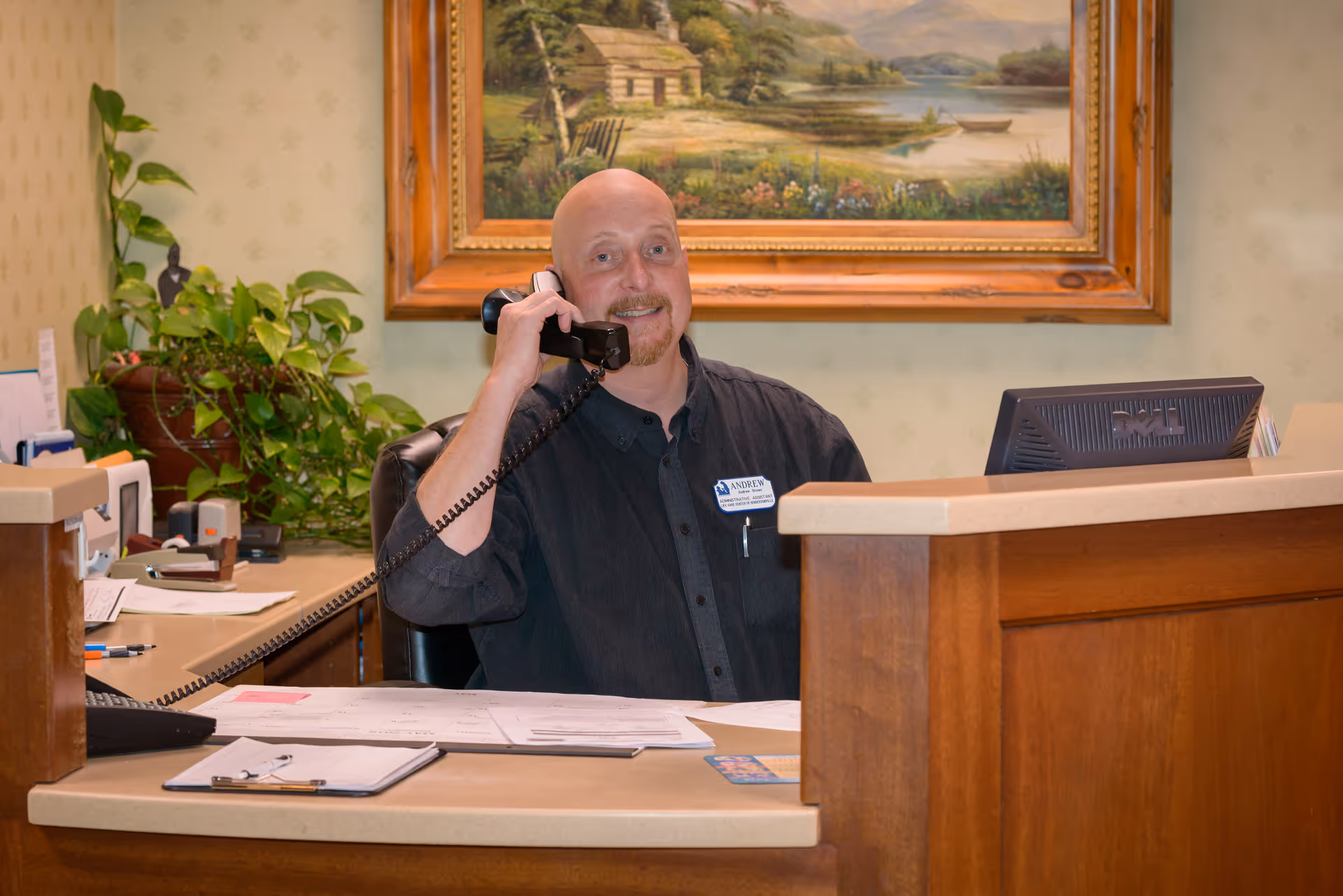 A man sitting behind a reception desk, smiling and talking on a corded telephone. The desk has papers, a clipboard, and a computer monitor. Behind him is a large framed painting of a countryside scene and a green leafy plant.