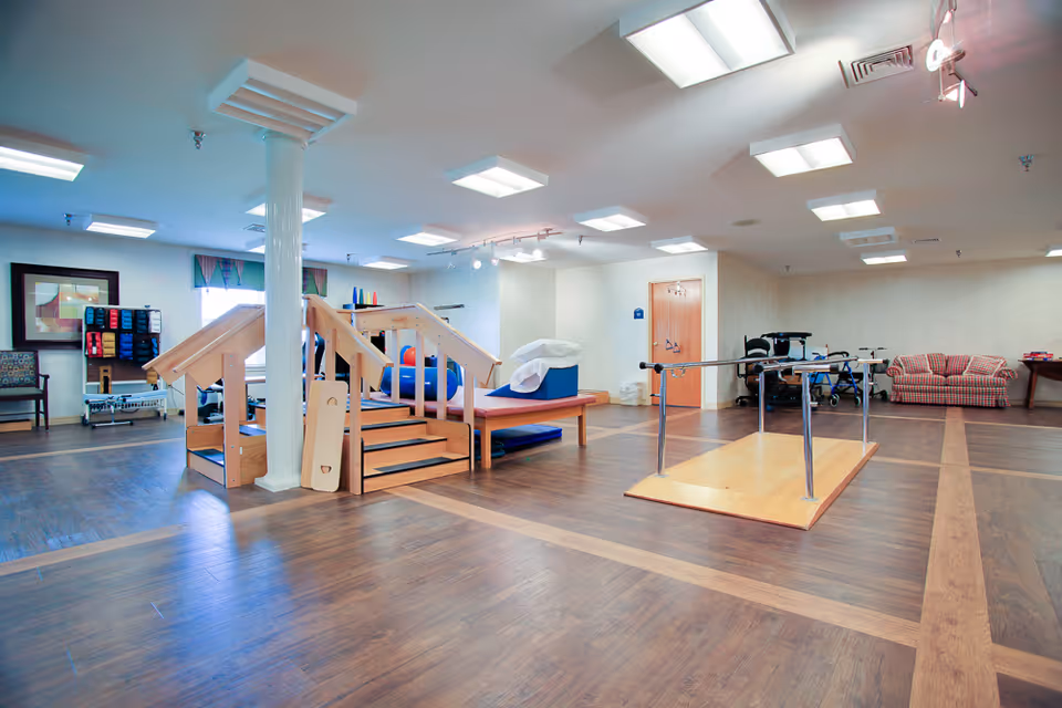 A spacious physical therapy room with wooden flooring, equipped with parallel bars for walking exercises, a small wooden staircase with handrails, therapy mats, exercise balls, and various rehabilitation equipment. There is a plaid couch and chairs along the walls, with bright overhead lighting and a framed picture on one wall.