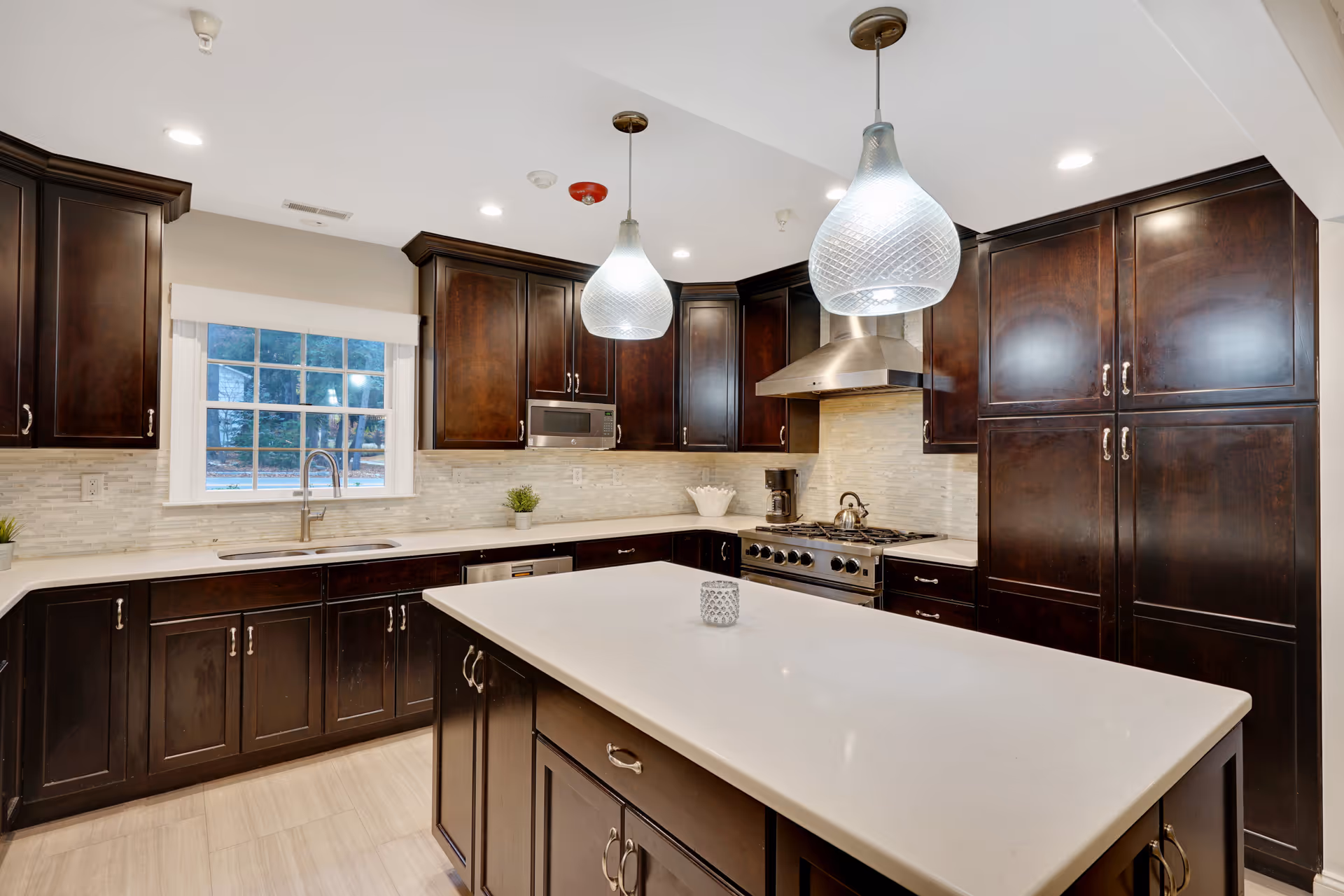 Modern kitchen with dark wood cabinets, a large white island countertop, stainless steel stove with hood, microwave, and two pendant lights hanging from the ceiling. There is a window above the sink showing an outdoor view.