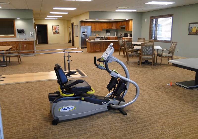 A spacious interior activity room with a NuStep recumbent exercise machine in the foreground, parallel bars, tables and chairs, and a kitchenette in the background.
