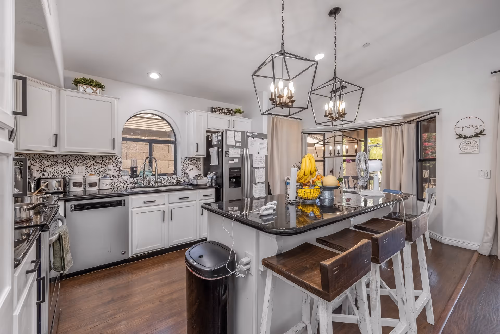 Bright kitchen with white cabinets, black countertops, and a central island with four wooden stools. Two modern pendant lights hang above the island. The kitchen features a stainless steel dishwasher, refrigerator covered with notes, and a window with an arched top. There is a fruit basket with bananas and other fruits on the island, and a sliding glass door with curtains leading to an outdoor area.
