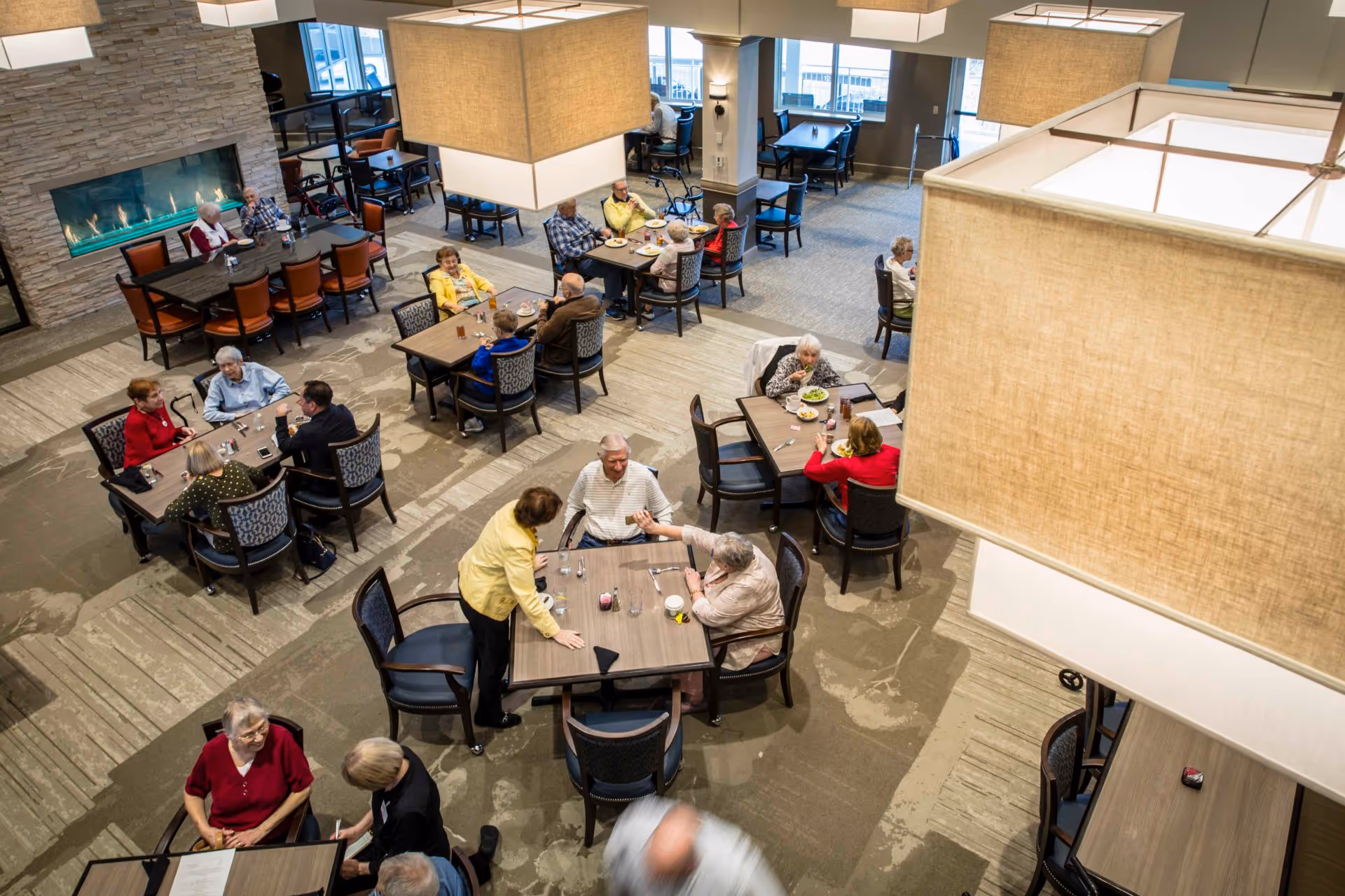 Bright assisted-living dining room with senior residents seated at multiple tables socializing and eating.
