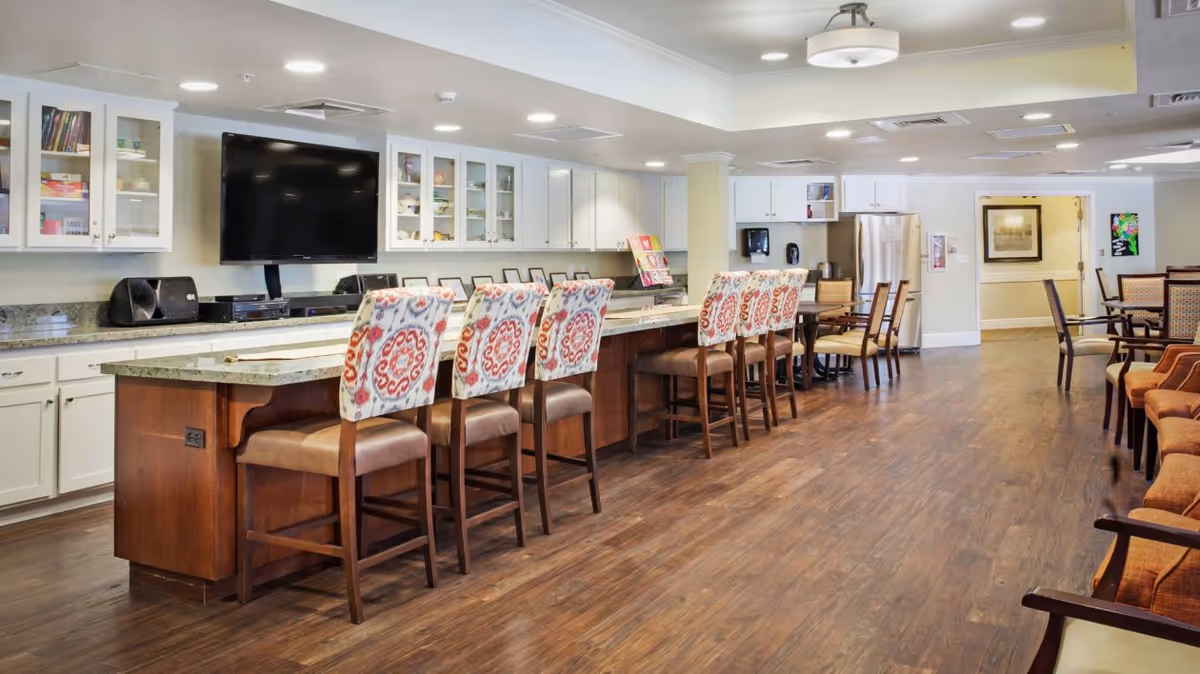 A spacious and well-lit common area with a long kitchen island featuring a granite countertop and several high chairs with patterned upholstery. The background includes white cabinets with glass doors, a large flat-screen TV mounted on the wall, a stainless steel refrigerator, and additional seating with tables and chairs arranged neatly on a wooden floor.