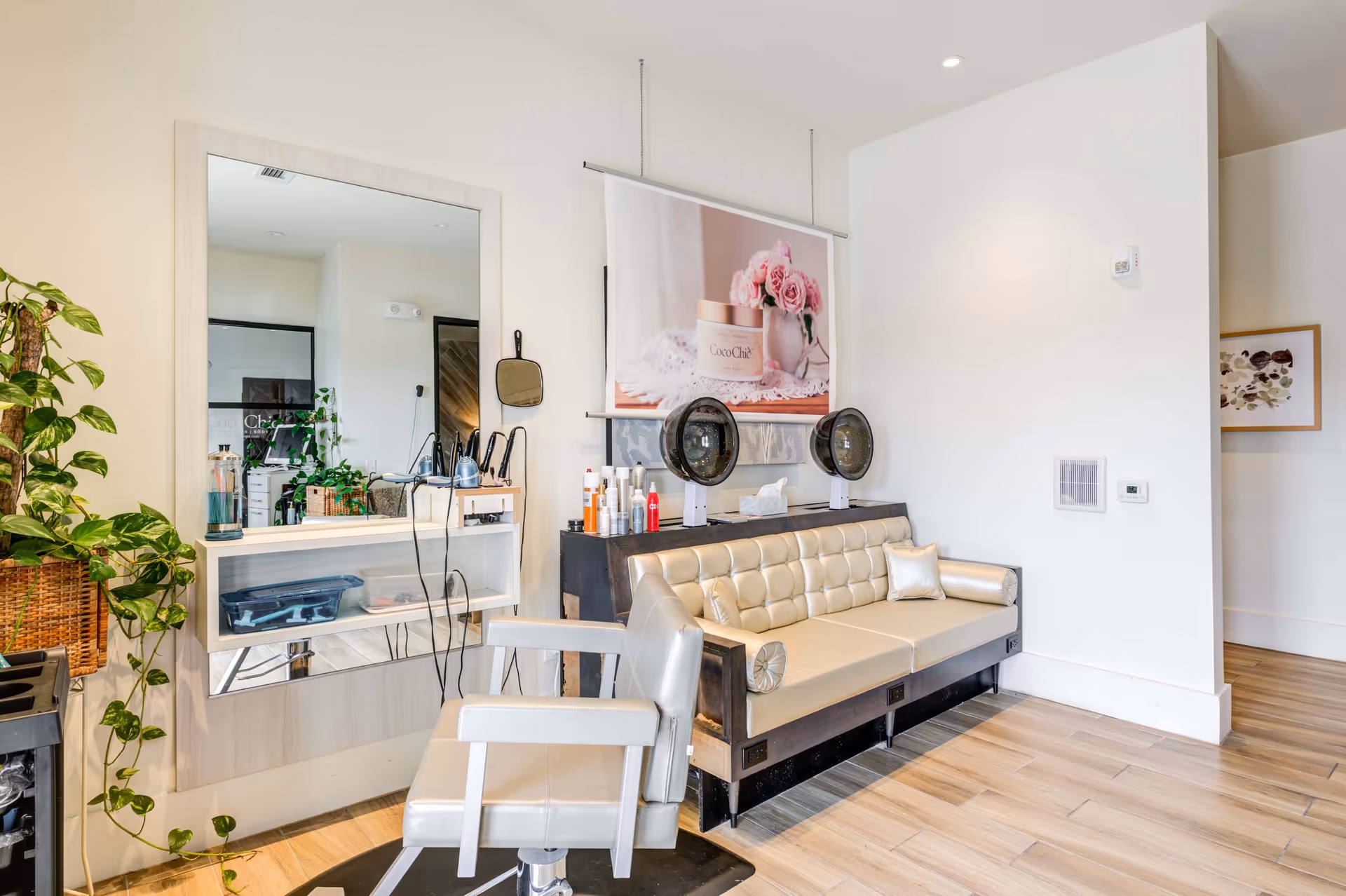 Bright salon-style interior with a styling station and mirror, a salon chair, two hooded hair dryers, and a tufted waiting sofa.