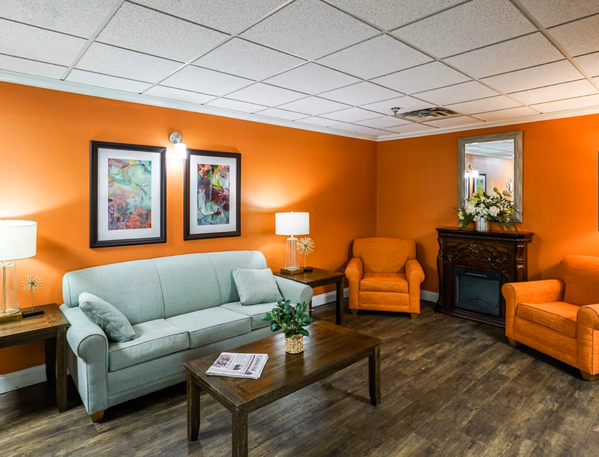 Common-area living room with a light blue sofa, orange armchairs, a wooden coffee table, and a decorative fireplace against orange walls.