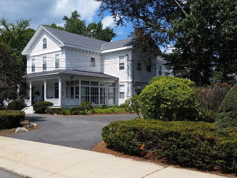 White two-story house-style senior living building with a covered porch, driveway, and landscaped shrubs under a partly cloudy sky.