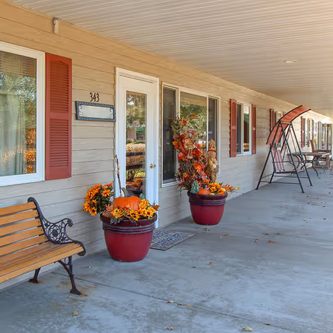 Covered front porch of a senior living facility with benches, potted autumn plants and pumpkins, entry doors and a swing.