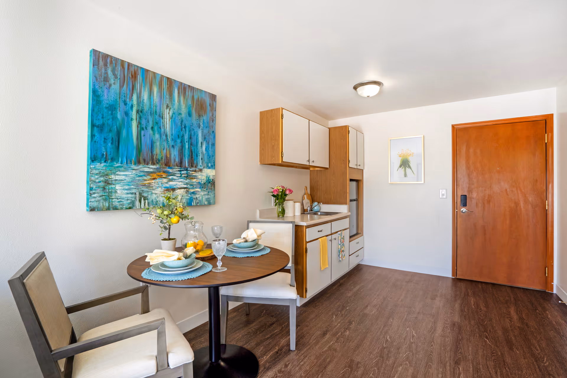 A small dining area with a round wooden table set for two with plates, napkins, and glasses. Two cushioned chairs are placed around the table. Behind the table is a kitchenette with wooden cabinets, a small sink, and a mini refrigerator. The room has wooden flooring, a wooden door, and two pieces of wall art, one large abstract painting and one smaller floral print.
