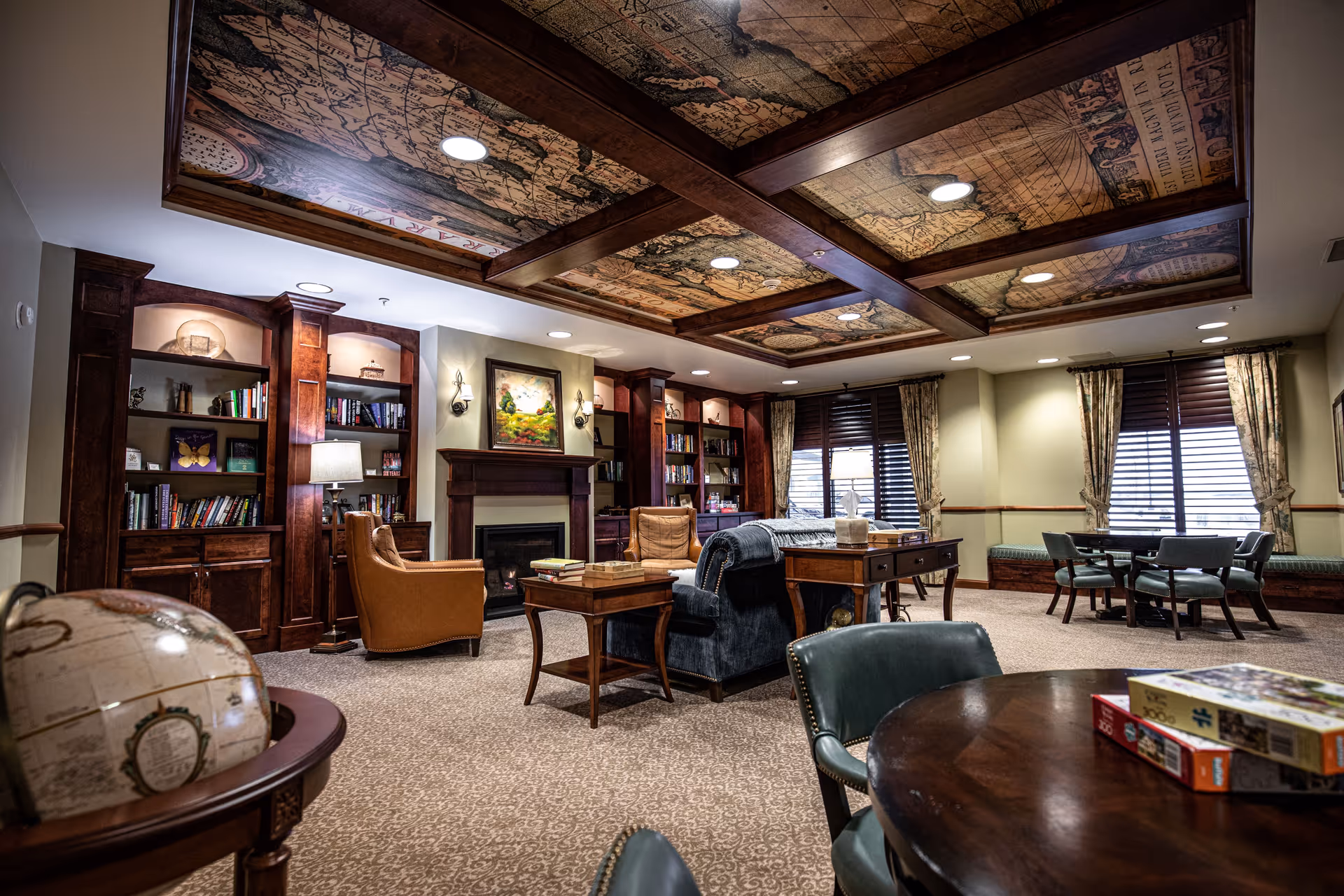 Cozy communal lounge with bookshelves, a fireplace, multiple seating areas, game tables, and a decorative coffered ceiling.