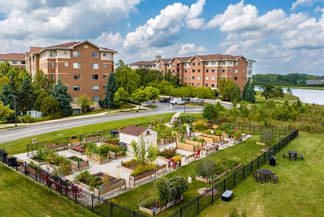 Aerial view of Sedgebrook's fenced community garden with raised beds in front of multi-story brick senior living buildings and a nearby lake.