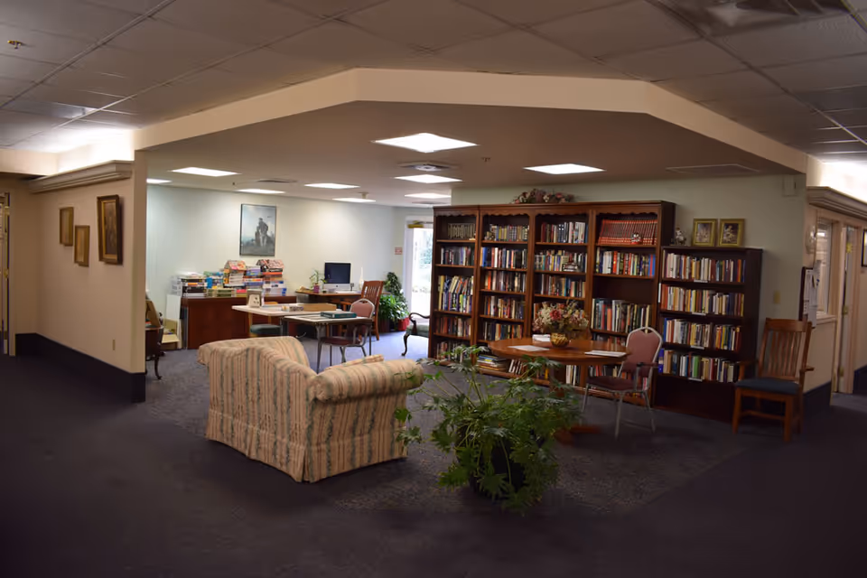 A comfortable communal library/lounge with bookshelves, tables, chairs, a patterned couch and potted plants.