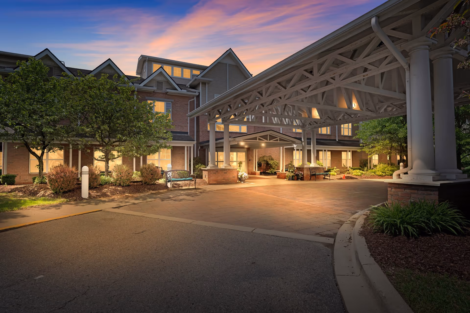 Exterior view of Town Village Sterling Heights at dusk, showing a covered entrance with white pillars and a brick building with lit windows surrounded by trees and landscaping.