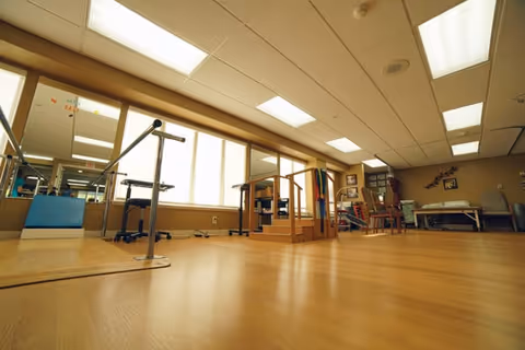 A spacious physical therapy or rehabilitation room with wooden flooring, parallel bars for walking exercises, a small staircase with handrails, chairs, and exercise equipment. Large windows allow natural light to fill the room, and mirrors line one wall.