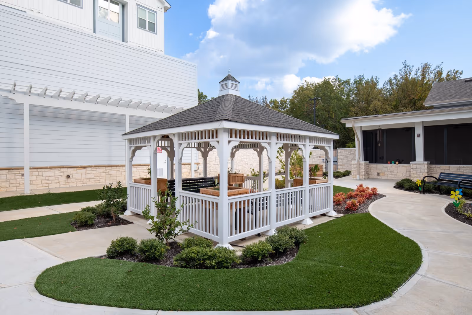 A white wooden gazebo with a shingled roof situated in a landscaped outdoor area with green grass, bushes, and paved walkways. The gazebo contains benches and planter boxes. Surrounding buildings with light-colored stone and siding are visible under a partly cloudy sky.