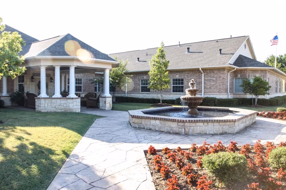 Outdoor view of Magnolia Assisted Living and Memory Care facility showing a stone-paved walkway leading to a covered seating area with white columns. There is a multi-tiered water fountain in the center surrounded by landscaping with red flowers and green shrubs. The building has brick walls and a sloped roof, with an American flag visible in the background.