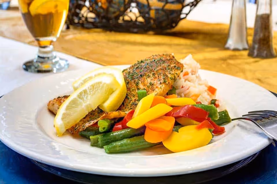 A white plate with a serving of seasoned fish topped with two lemon wedges, accompanied by mixed steamed vegetables including green beans, carrots, yellow squash, and red bell peppers, and a side of rice with diced vegetables. A glass of iced tea and salt and pepper shakers are visible in the background on a yellow tablecloth.