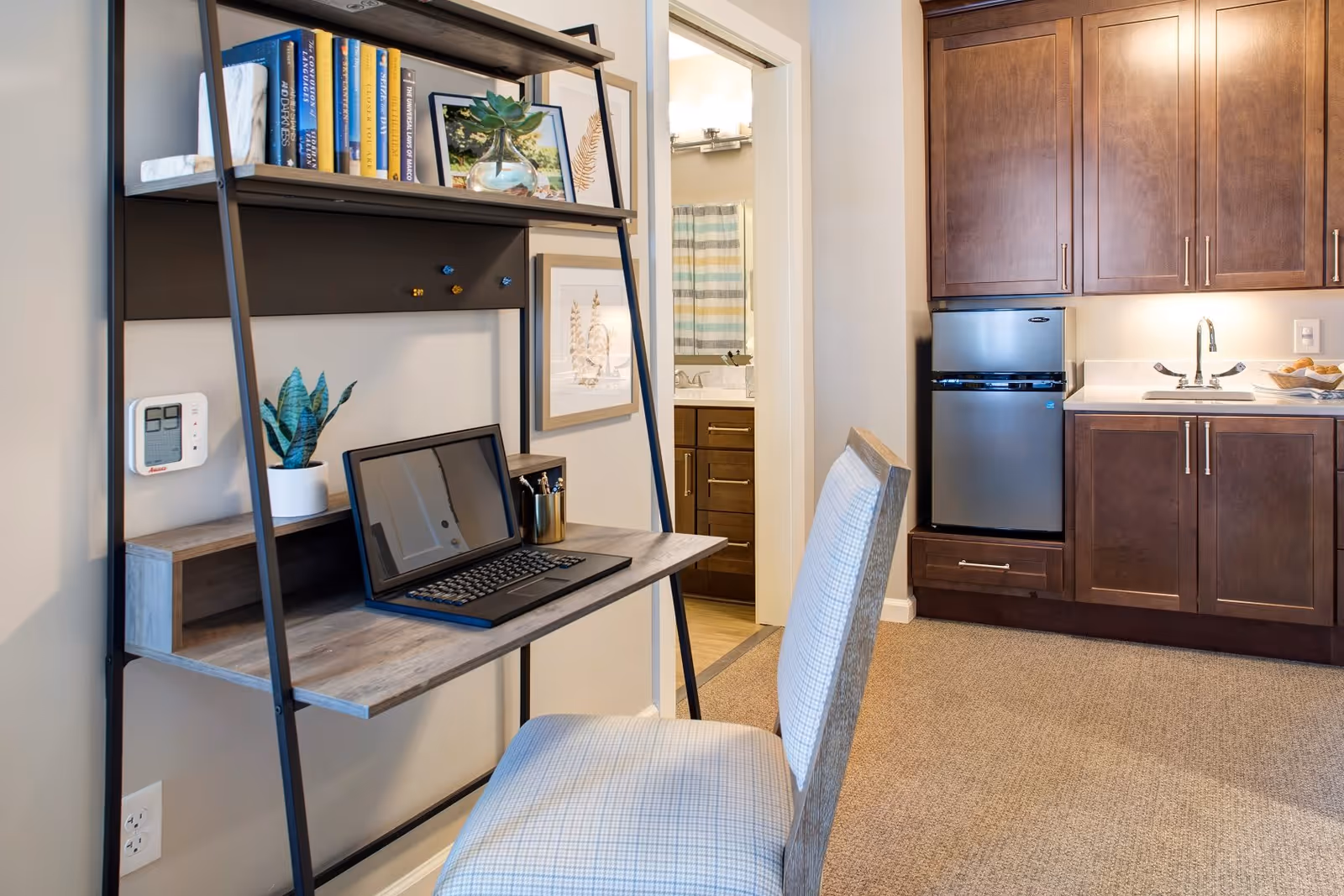 A cozy interior space featuring a small desk with a laptop, books, and a potted plant. A cushioned chair is positioned in front of the desk. To the right, there is a kitchenette area with dark wooden cabinets, a small refrigerator, and a sink. In the background, an open door reveals a bathroom with a vanity, mirror, and a striped shower curtain.