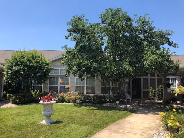 Exterior view of a single-story building with beige siding and a brown roof, surrounded by green trees, shrubs, and a well-maintained lawn. A concrete walkway leads to the entrance, and a decorative white planter with flowers is placed on the grass.