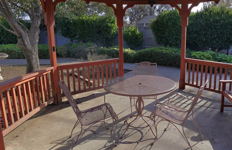 Outdoor covered patio area with a round metal table and four matching metal chairs. The patio is surrounded by a wooden railing and overlooks a garden with bushes and trees.