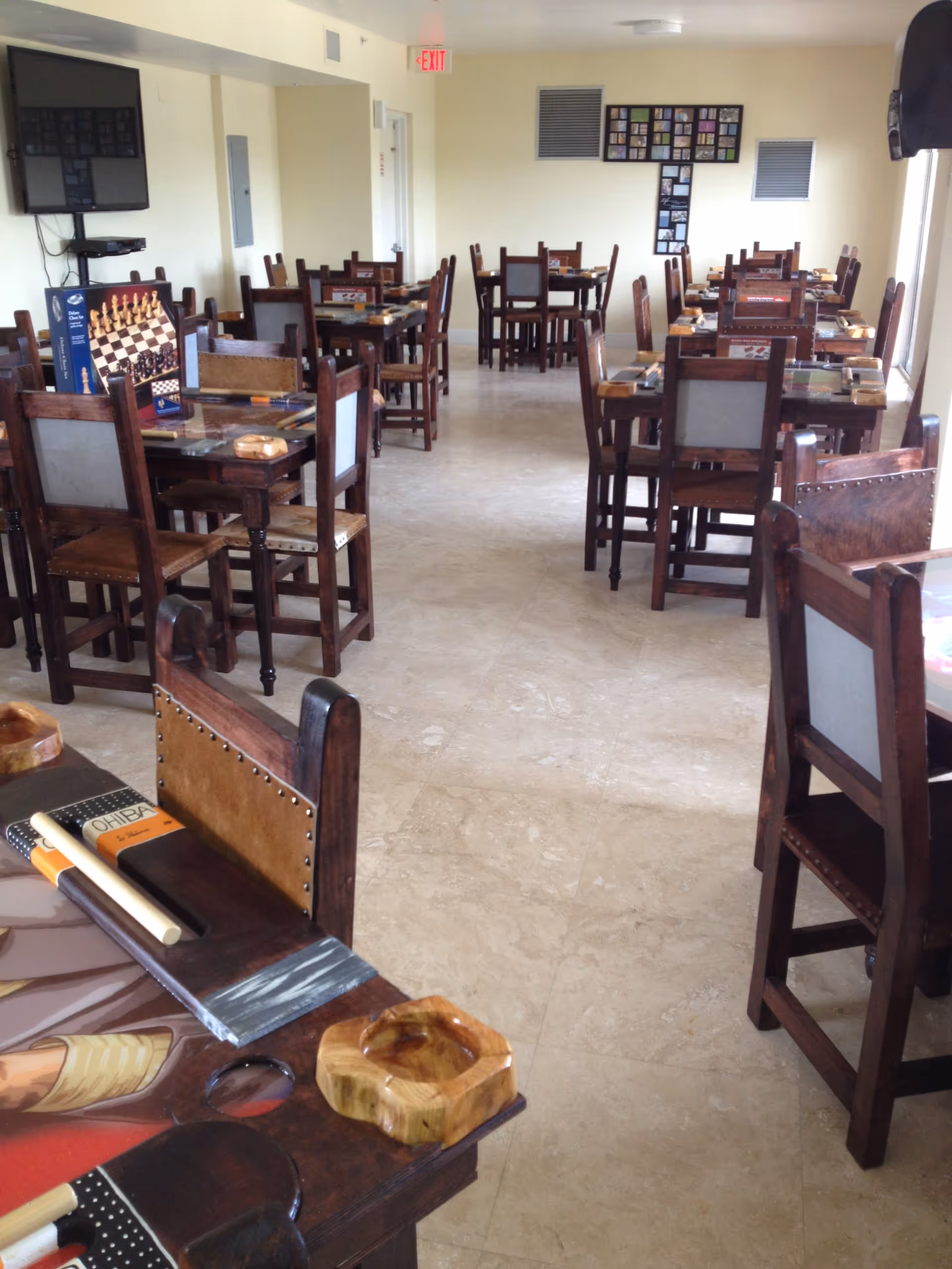 Interior view of a room with multiple wooden tables and chairs arranged neatly. Each table has a chessboard and some game pieces on it. The room has a tiled floor, light-colored walls, a wall-mounted TV, and a framed collage on the far wall. An exit sign is visible above a door in the background.