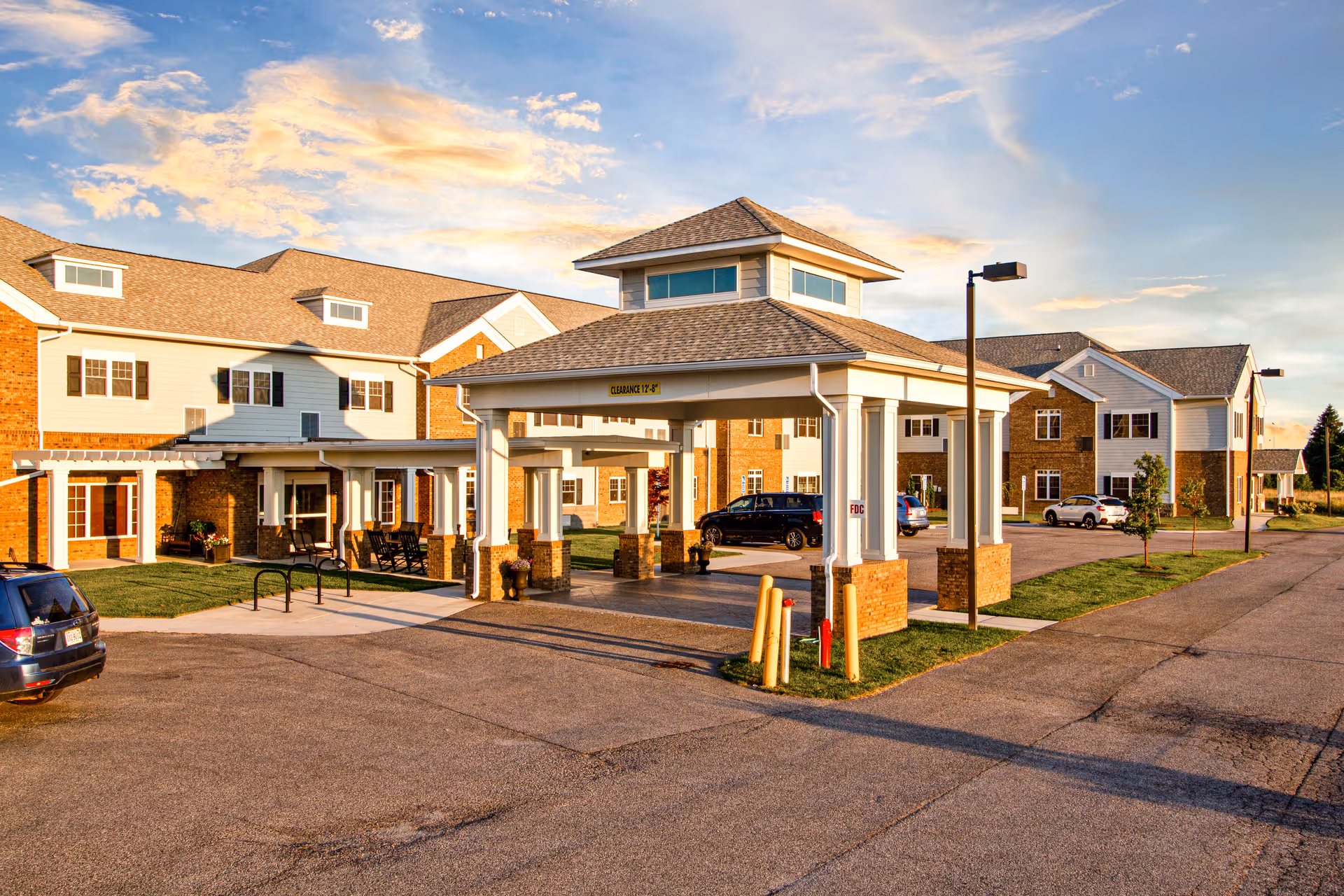 Exterior view of English Meadows Blacksburg Campus showing a covered entrance with a clearance sign, multiple parked cars, and a multi-story building with brick and siding under a partly cloudy sky at sunset.