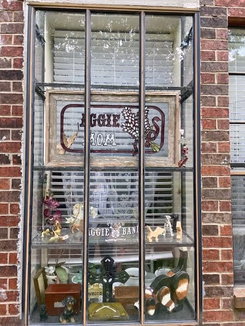 Exterior view of a brick building window displaying shelves of small decorative figurines and a framed sign behind blinds.