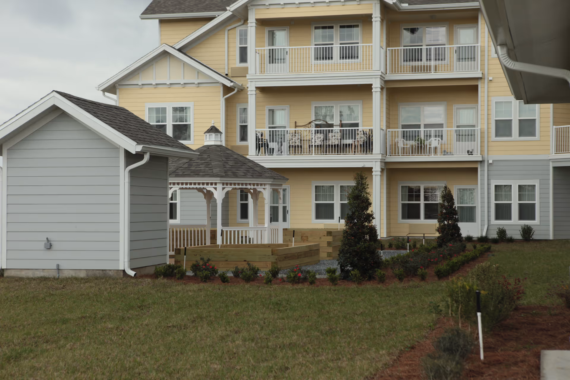 View of the exterior of a multi-story residential building with yellow and gray siding, featuring balconies with outdoor furniture. In the foreground, there is a small white gazebo, a gray shed, landscaped garden beds with shrubs and flowers, and a grassy lawn.
