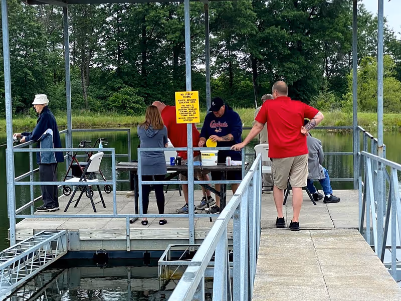 A group of people standing and sitting on a metal dock over a pond surrounded by trees. One person is fishing, others are gathered around a table with fishing supplies. The dock has railings and a sign with fishing rules.