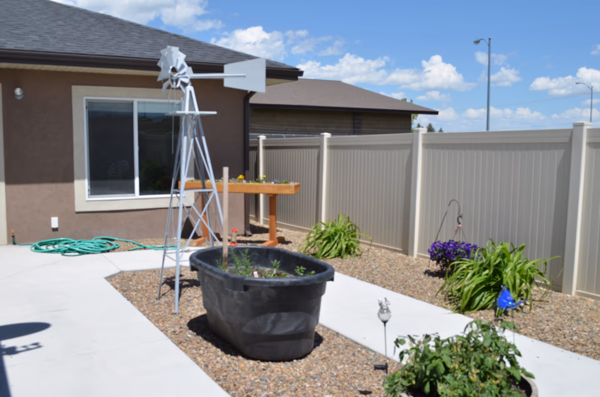 Outdoor garden area with a small windmill decoration, a black planter tub with plants, a raised wooden planter box, green plants, and a beige privacy fence under a blue sky with scattered clouds.