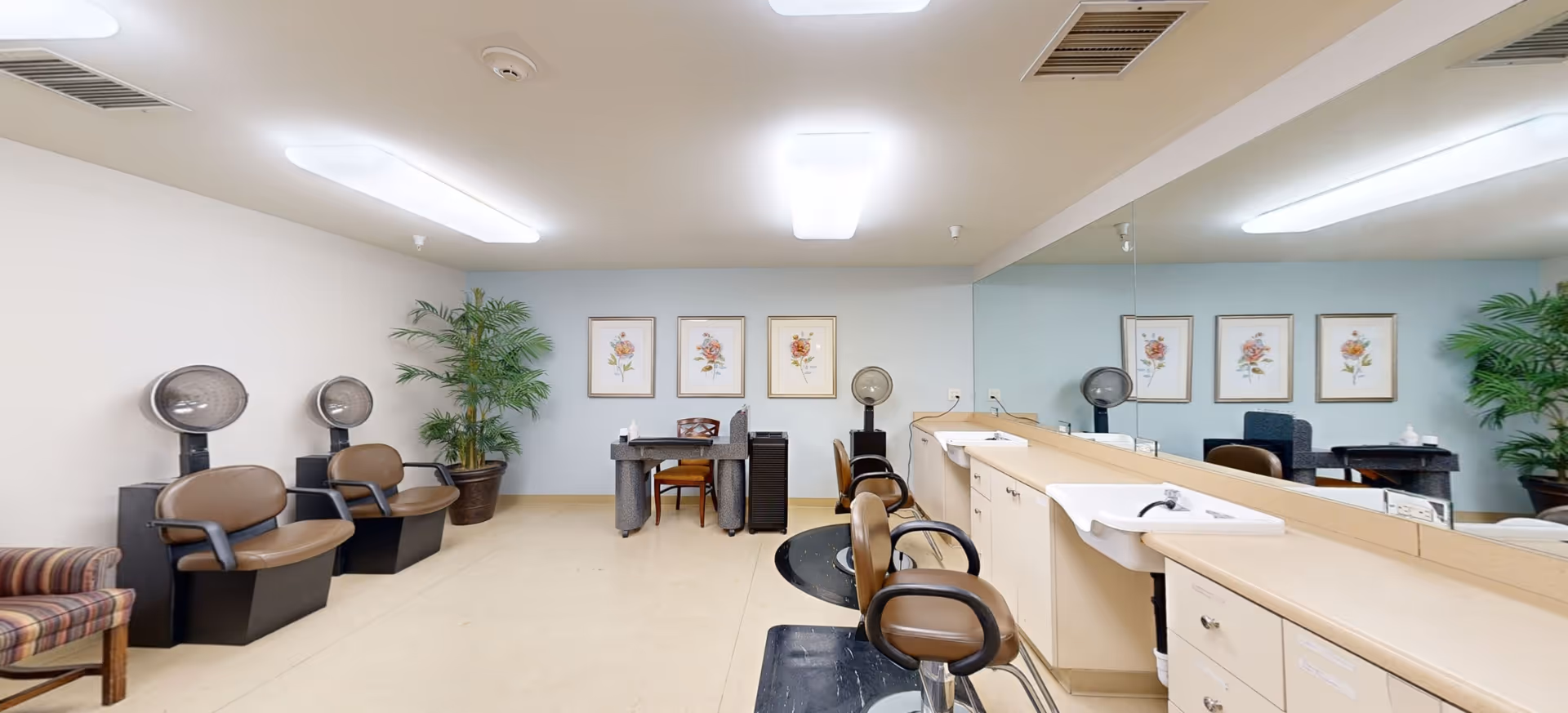Interior view of a salon area in a senior living facility with multiple salon chairs, hair dryers, a long counter with sinks, and a large mirror. There are framed floral pictures on the wall and potted plants in the corners.