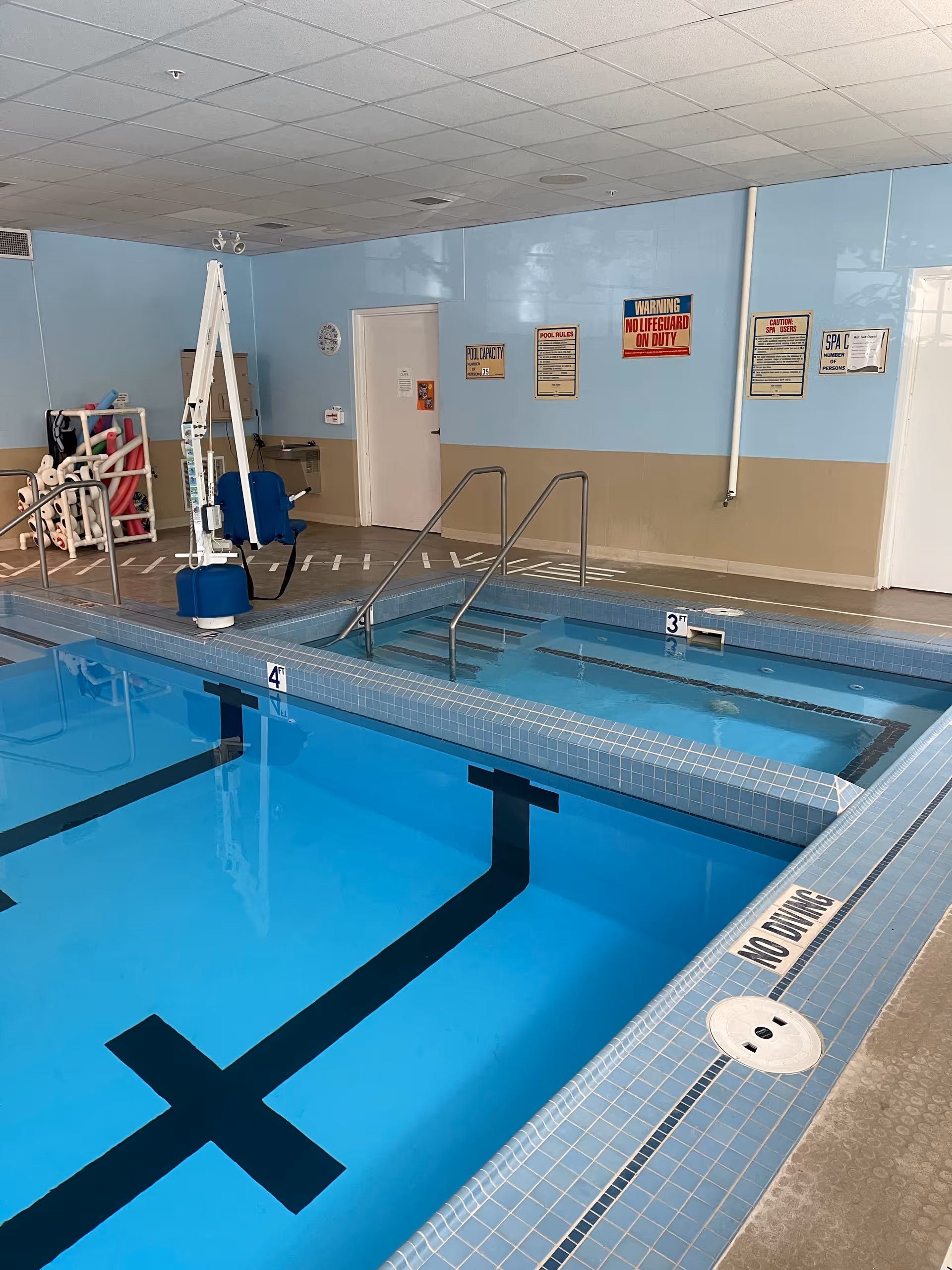 Indoor swimming pool area with a pool lift, metal handrails, depth markers, and safety signs on the wall.