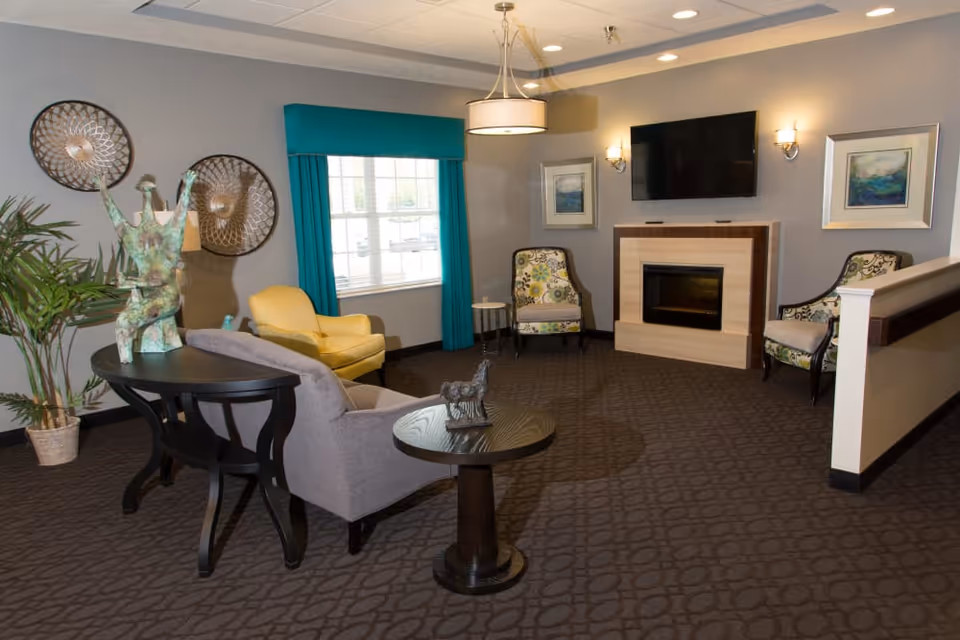 Furnished lounge with chairs, a sofa, round tables, a fireplace with a TV above, and a window with teal curtains.