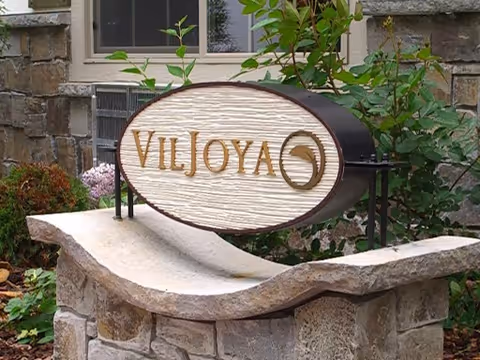 Stone sign with the name 'Viljoya' mounted on a curved stone base surrounded by plants and greenery, with a building wall and window in the background.