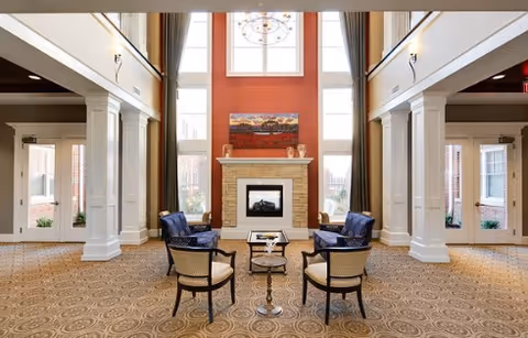 A spacious and elegant living area with high ceilings, large windows, and a central fireplace. The room features four chairs arranged around a small table on a patterned carpet, with tall white columns and warm lighting.