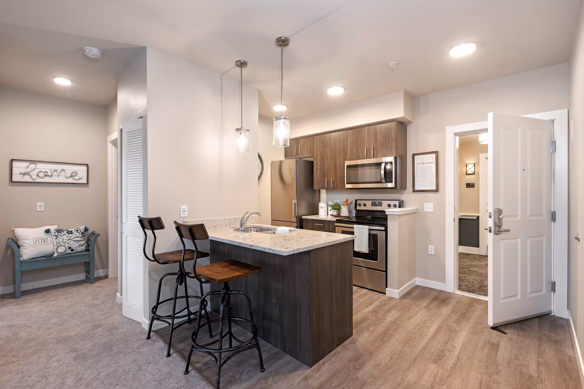 Open-plan apartment kitchen with a granite island and three bar stools, stainless steel appliances, and an open entry door to a hallway.
