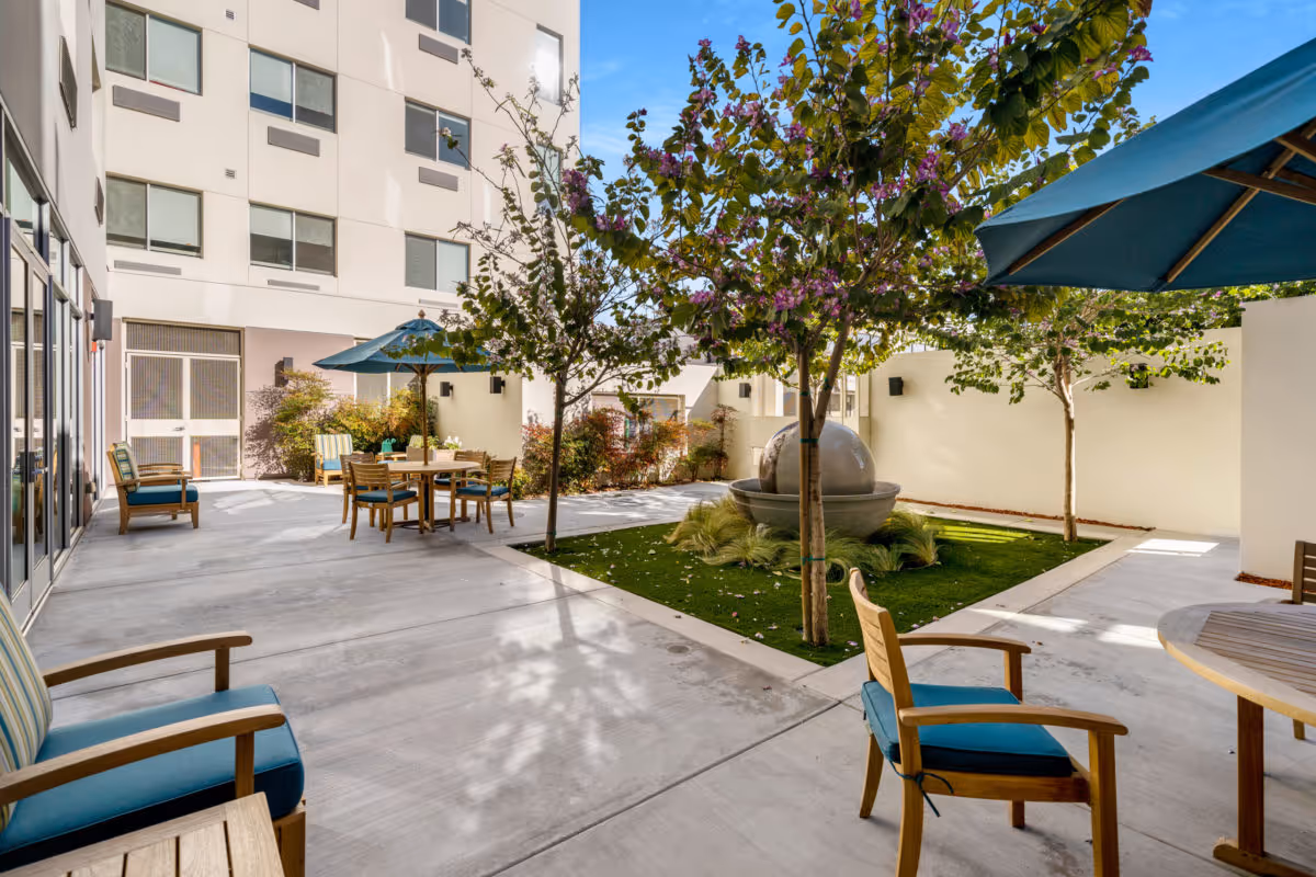 Outdoor courtyard area at a senior living facility with several wooden chairs with blue cushions, round tables with blue umbrellas, small trees, and a central water fountain surrounded by greenery. The courtyard is enclosed by a white building and walls.