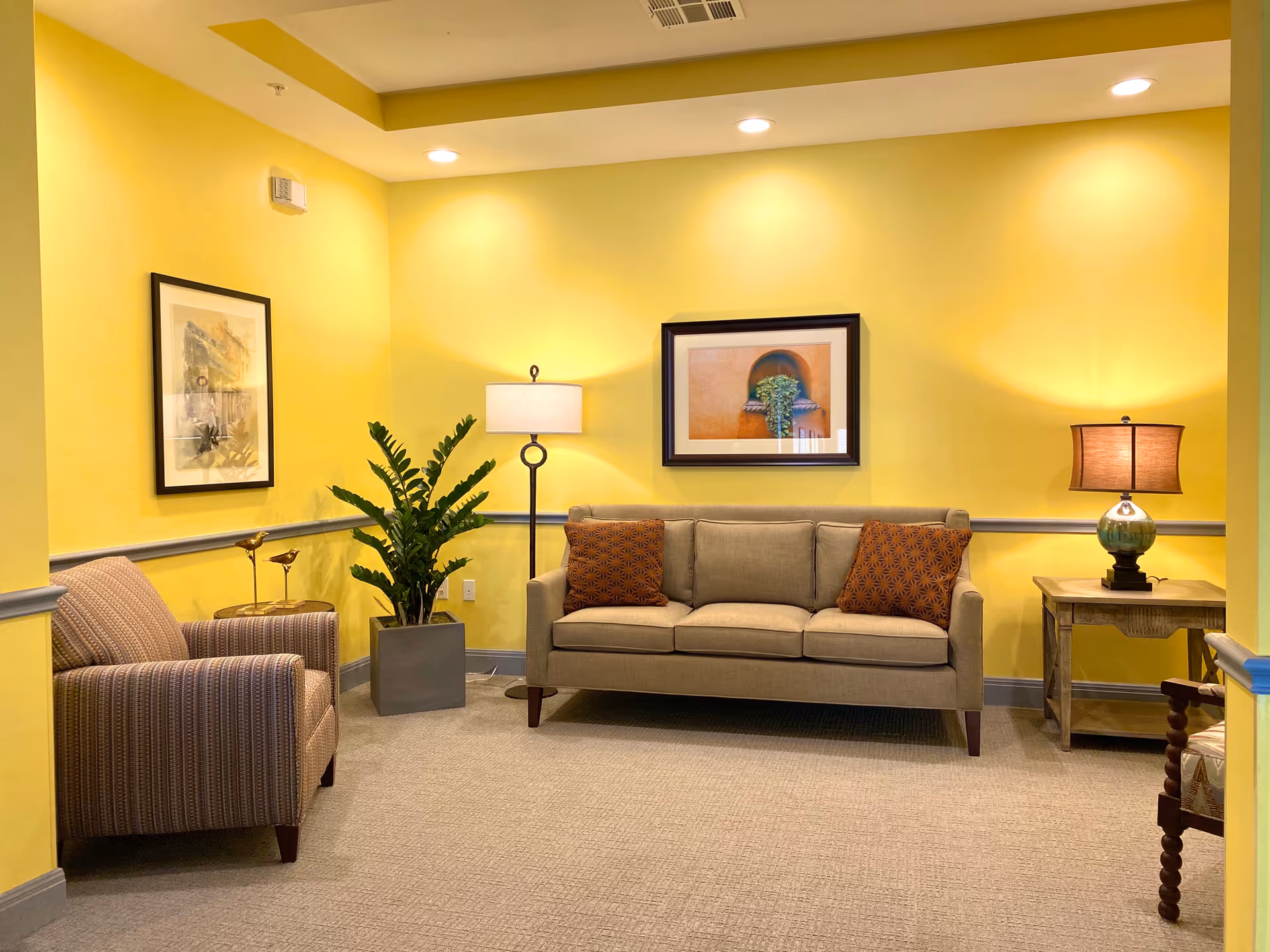 A cozy living room with bright yellow walls and gray trim. The room features a beige sofa with two patterned orange pillows, a striped armchair, a wooden side table with a lamp, a floor lamp, a potted plant, and two framed pictures on the walls. The floor is carpeted in a neutral tone.