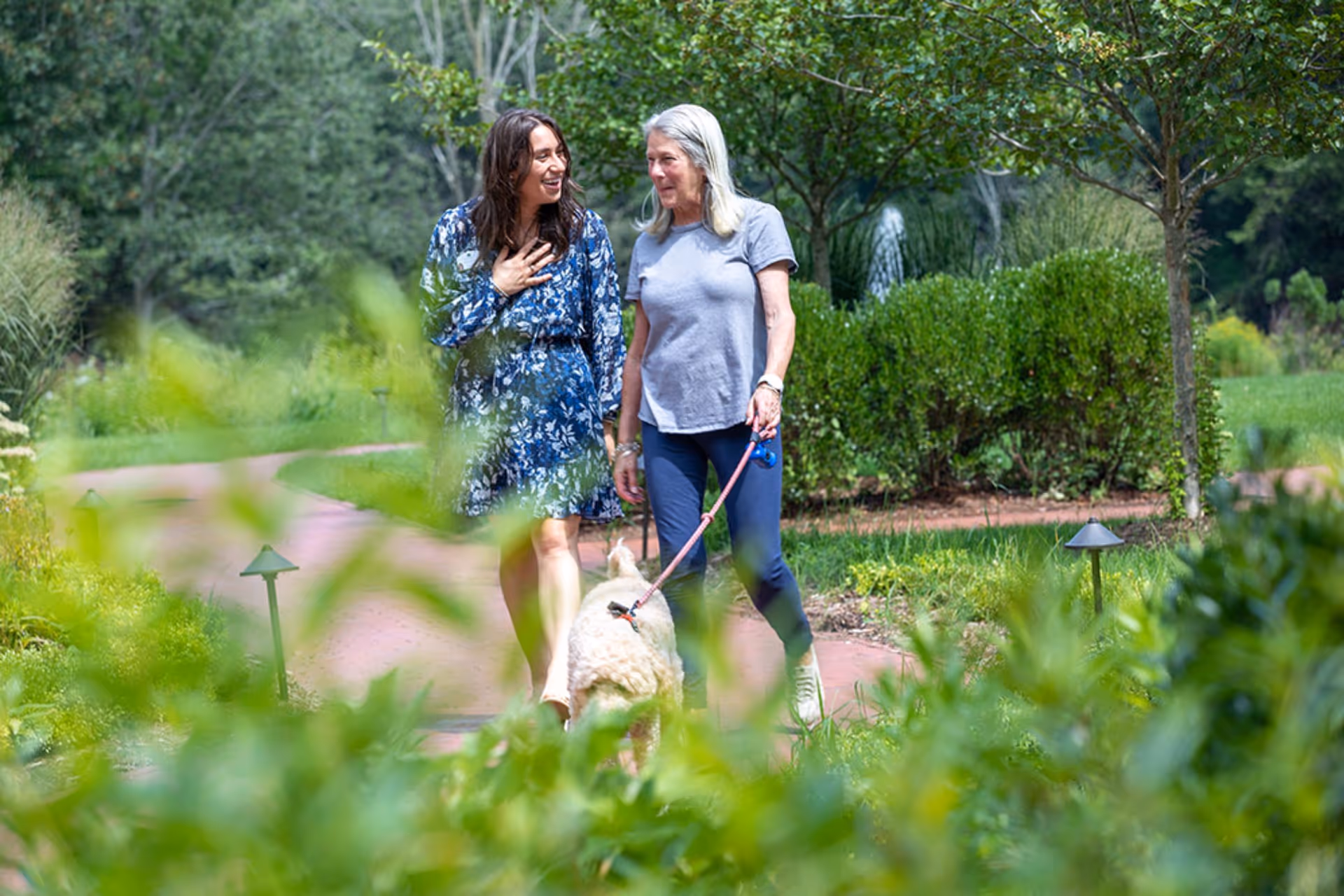 Two women walking and talking on a garden path surrounded by greenery, with one woman holding a dog on a leash.