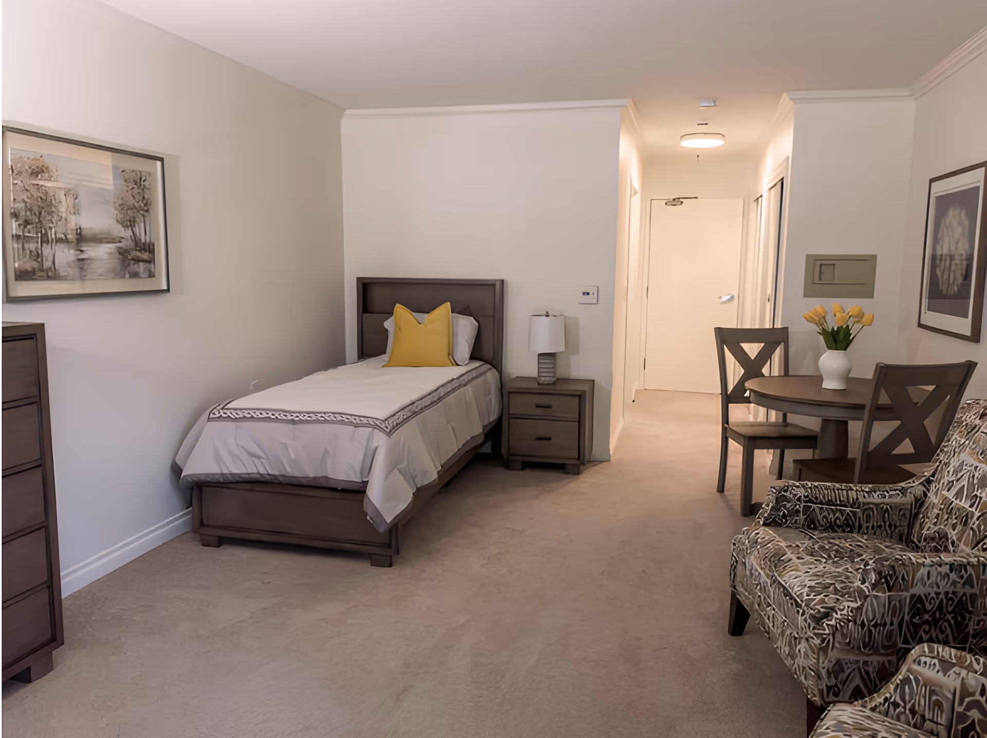 A neutral-toned studio bedroom with a single bed, nightstand, small dining table, and upholstered chairs.