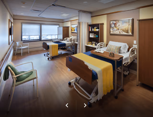 A spacious and well-lit hospital room with two patient beds, each equipped with a yellow blanket and a bedside table. The room has wooden flooring, a large window with blinds, a green chair with a blanket draped over it, and a bookshelf with decorative items and pictures. There is a curtain divider between the beds and framed artwork on the walls.