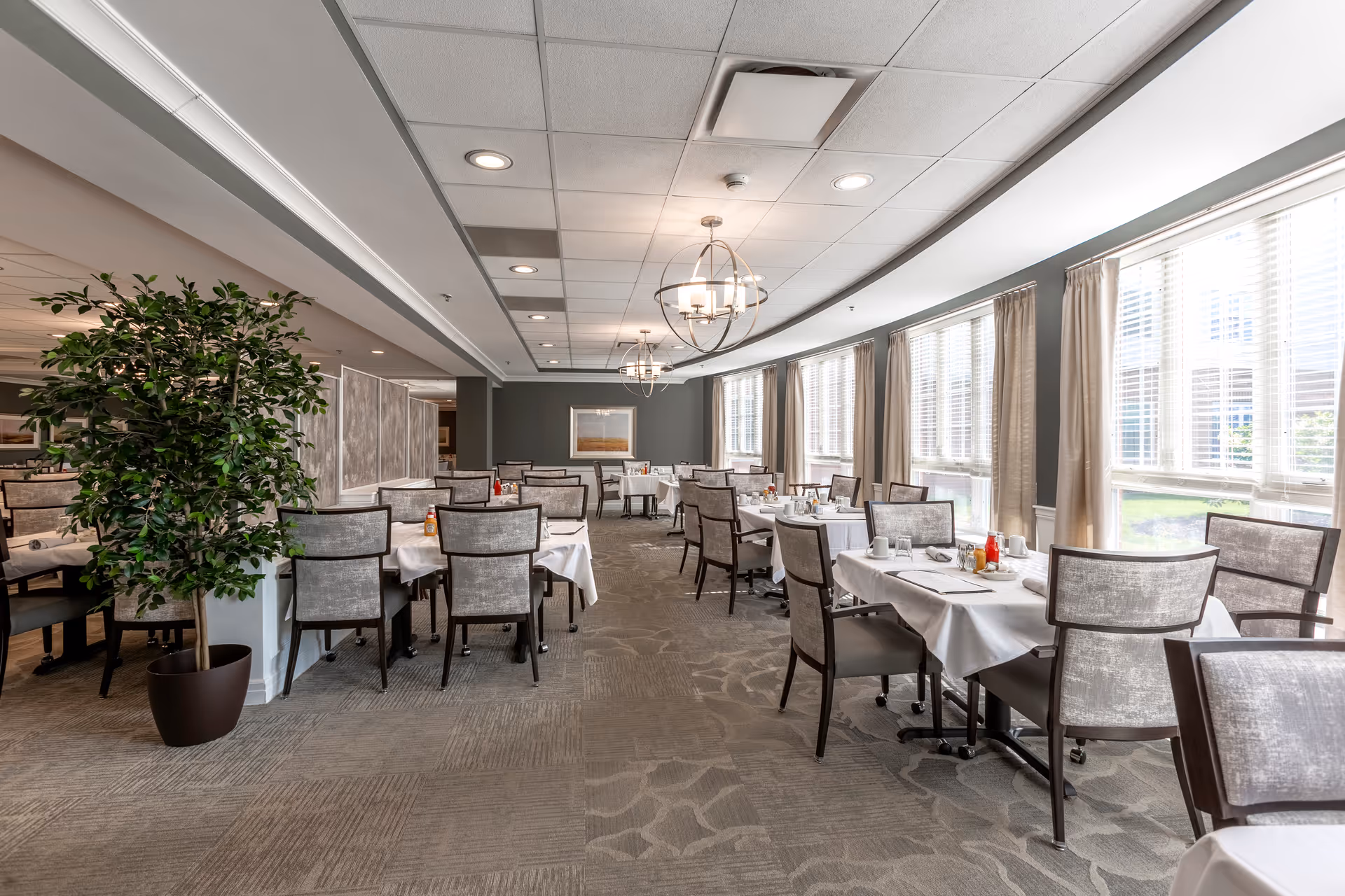 A bright dining room with multiple tables covered in white tablecloths, each set with glasses, plates, and condiments. The room features large windows with beige curtains allowing natural light to fill the space. There are modern chandeliers hanging from the ceiling and a large potted plant on the left side.