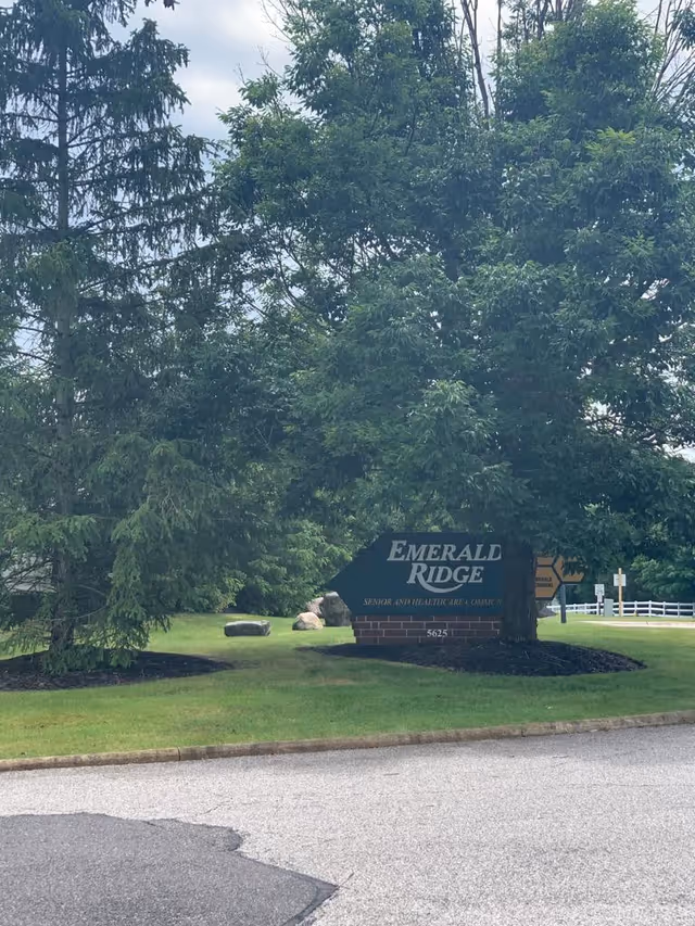 A green and brick sign for Emerald Ridge Senior and Healthcare Community partially obscured by a large tree, situated on a grassy area next to a paved road with other trees and a white fence in the background.