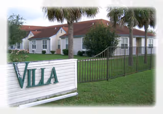Single-story light-colored assisted living building with palm trees, a lawn, a fence, and a white sign reading 'VILLA' in the foreground.