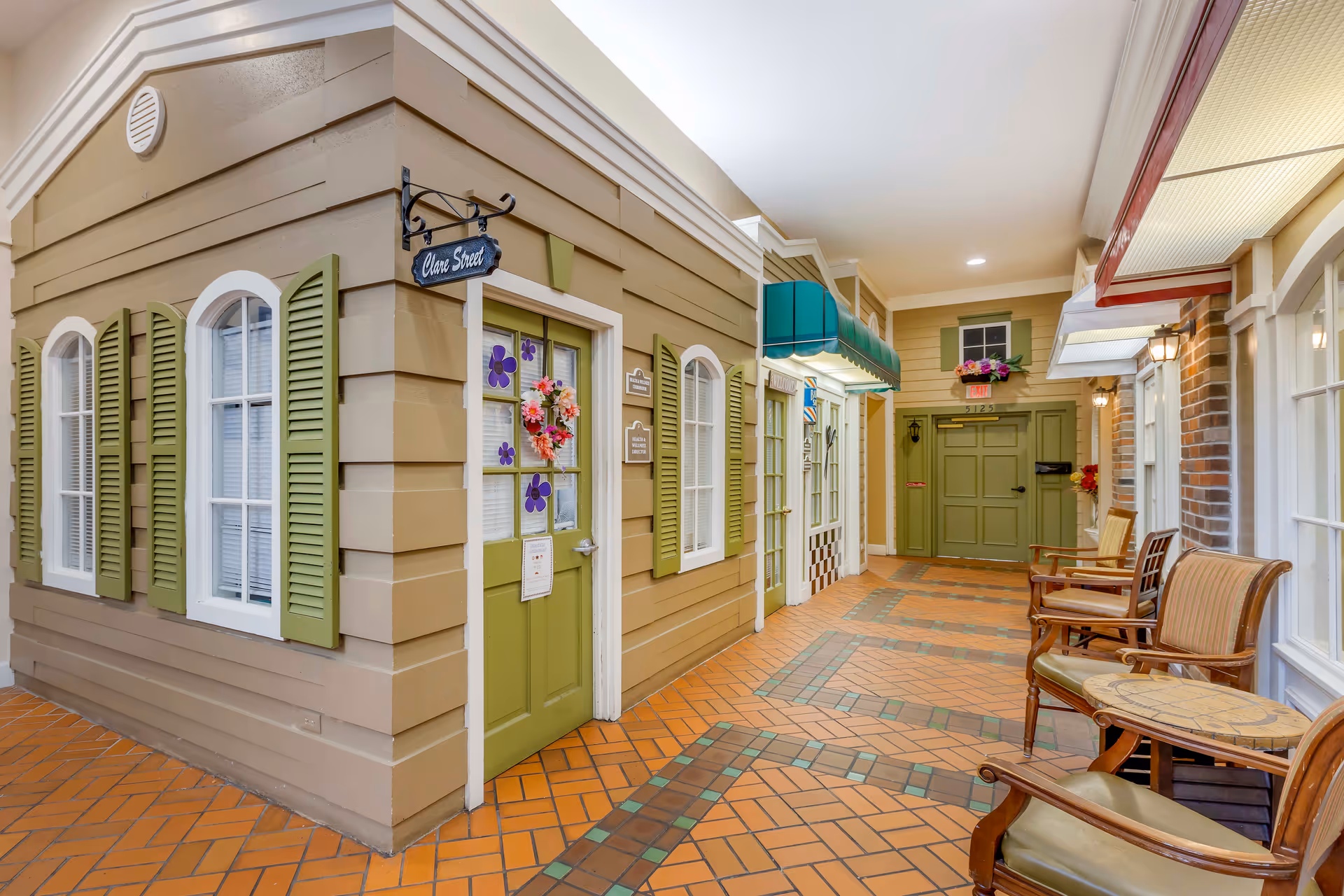 Indoor hallway designed to look like an outdoor street with building facades, green doors and shutters, decorative awnings, and a tiled floor. There are chairs and small tables along the right side, with warm lighting and floral decorations on some doors.