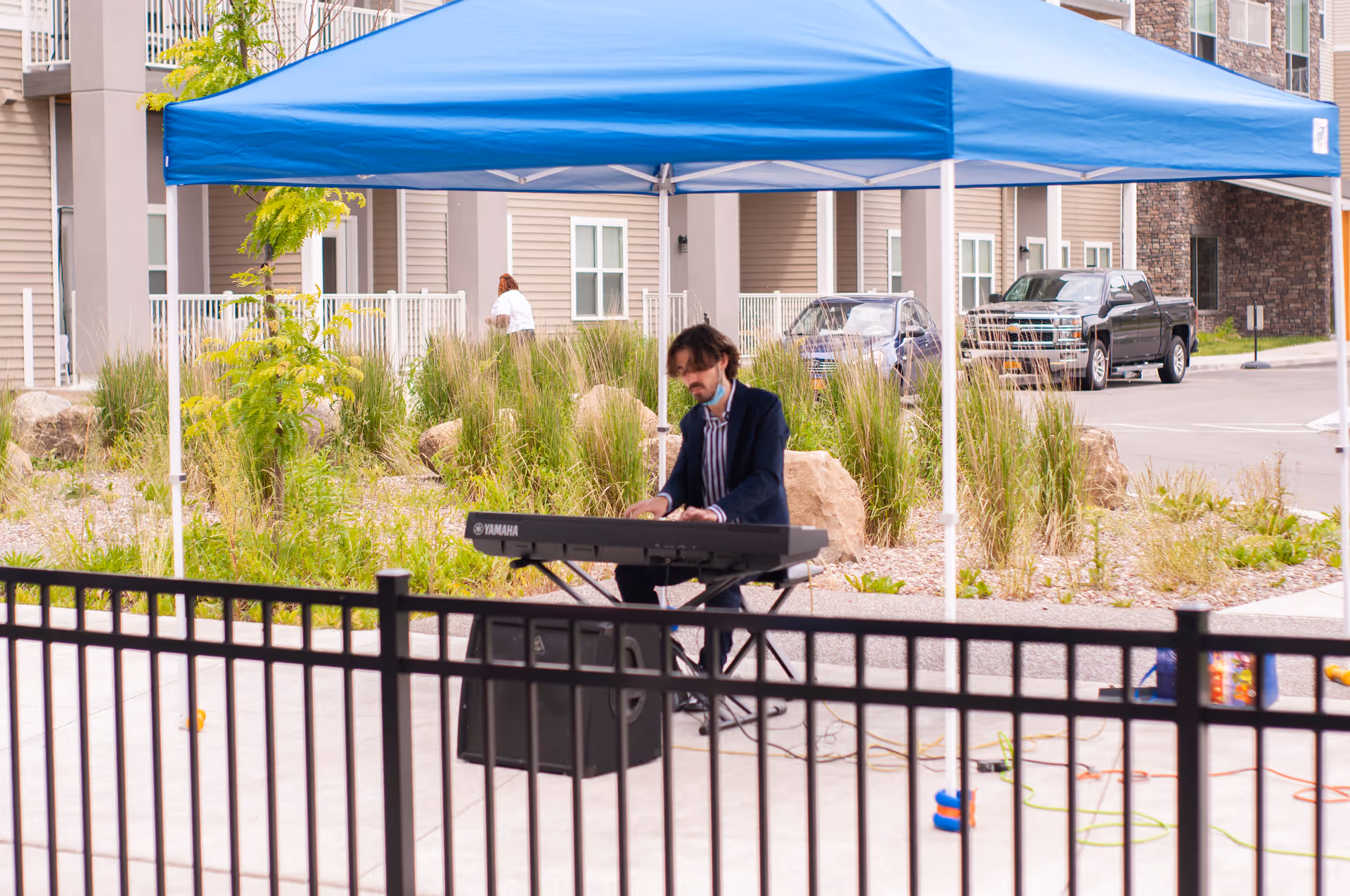 A musician plays an electronic keyboard under a blue pop-up canopy outside a senior living building.