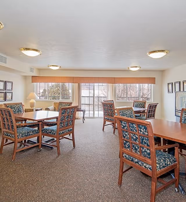 A bright and spacious common area with multiple wooden tables and chairs featuring blue patterned cushions. Large windows and sliding glass doors allow natural light to fill the room, with beige walls and carpeted flooring. There are framed pictures on the walls and a lamp on a side table near the windows.