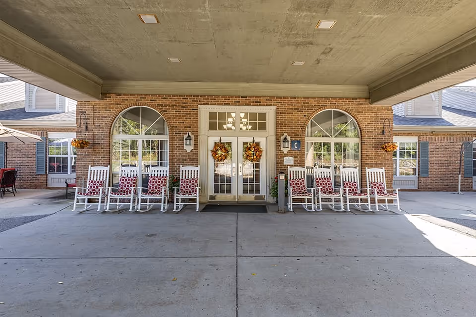 Covered brick entrance with double glass doors, white rocking chairs with patterned cushions, and autumn wreaths.