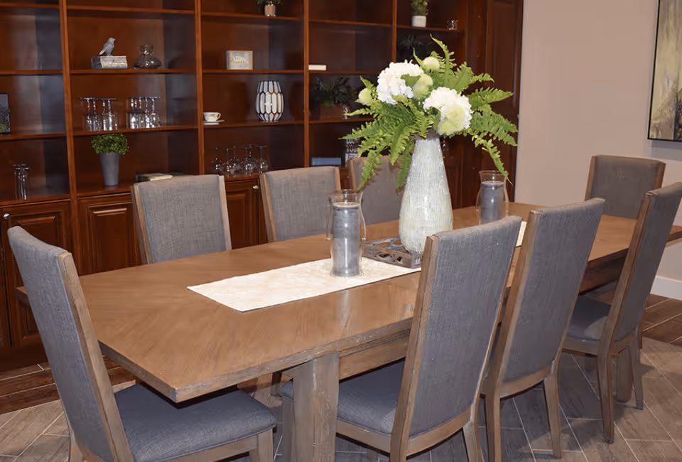 A dining room with a long wooden table surrounded by eight gray upholstered chairs. The table has a white runner and a large vase with white flowers and green ferns as a centerpiece. In the background, there is a wooden shelving unit with various decorative items including glassware, plants, and small ornaments.