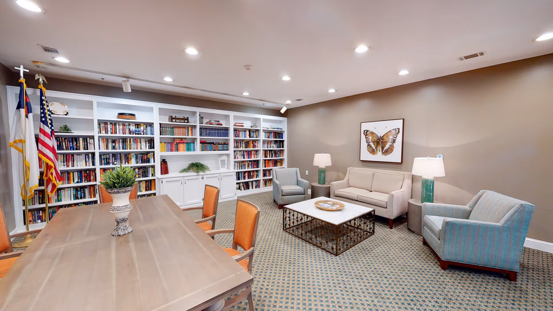 A cozy interior room featuring a large wooden table with orange chairs on one side and a seating area with a beige sofa, two blue armchairs, and a white coffee table. The back wall has built-in white bookshelves filled with books and decorative items. Two flags, including an American flag, stand in the corner. The room is well-lit with recessed ceiling lights and has a framed butterfly artwork on the wall.