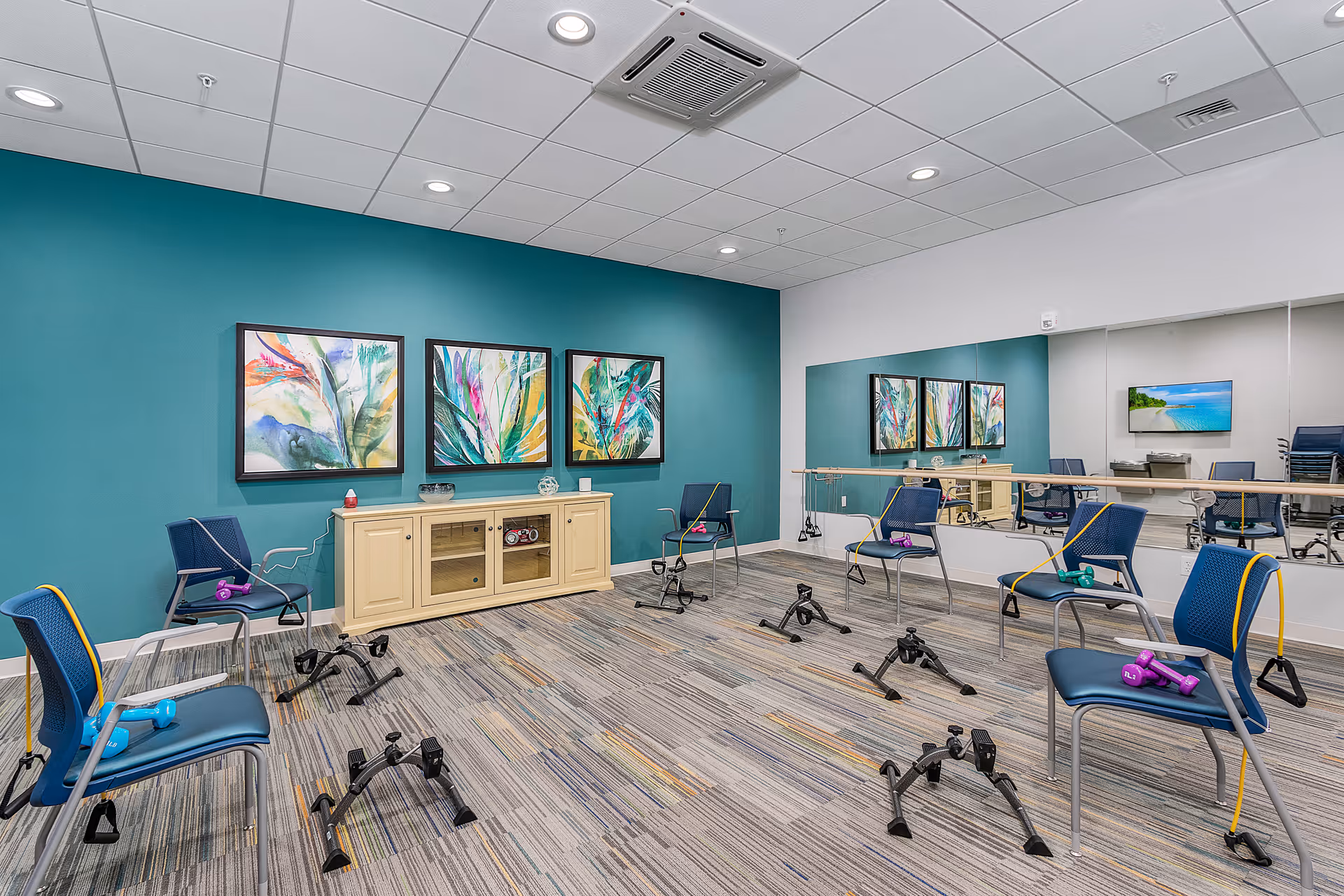 A bright exercise room with six blue chairs arranged in a circle, each equipped with resistance bands and small dumbbells. The room has a teal accent wall adorned with three colorful abstract paintings and a cream-colored cabinet beneath them. A large mirror covers one wall, reflecting the room and a TV mounted on the opposite wall. The floor is carpeted with a patterned design, and the ceiling has recessed lighting.